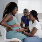 A pregnant woman sits in a chair looking down while a doula feels her belly, and a man sits in a chair to the side, watching them.